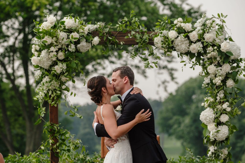 Bride and groom share a first kiss surrounded by why flowers at greenery.