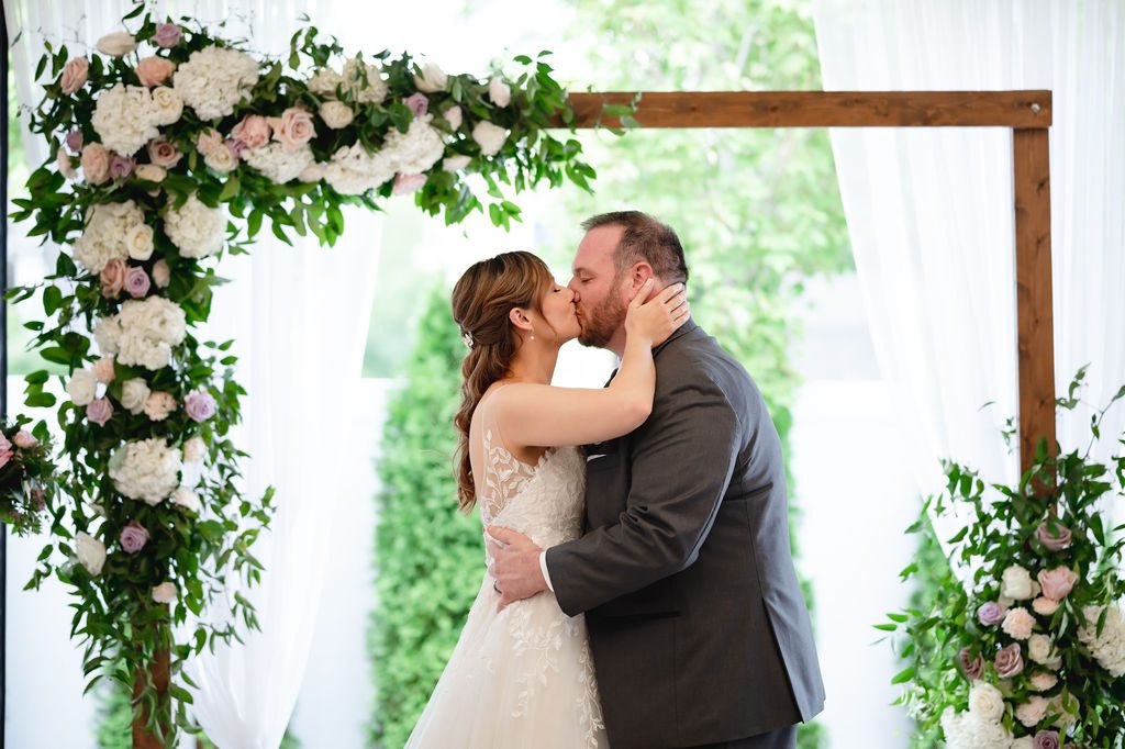 Bride and groom share their first kiss in front of a square arch.