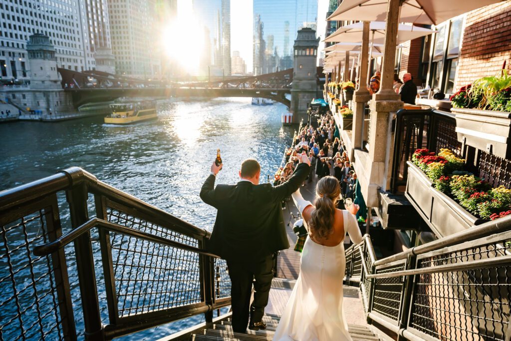 Wedding guests cheer on bride and groom as they walk down the stairs outside at River Roast in Chicago, IL.